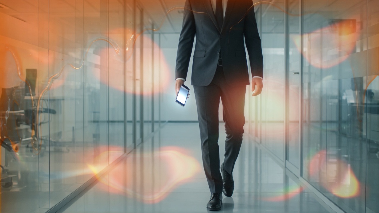 An adult walking a glass office corridor with a phone in hand